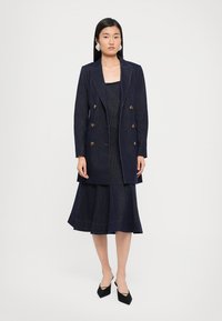 Marineblauwe spijkerjurk met een getailleerde lijfje en een uitlopende rok, gecombineerd met een bijpassende dubbelzijdige blazer met gouden knopen.