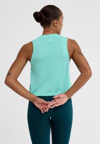 Helles Türkis ärmelloses Oberteil aus weichem Material, mit einem cropped Design, glatter Textur und schlichter runder Ausschnitt. Getragen zu dunklen Leggings.