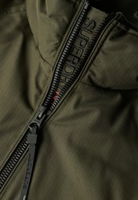 Olivgrüne Zip-Jacke aus strukturiertem Stoff mit Karomuster, schwarzer Reißverschluss mit "SUPERDRY"-Branding und Stehkragen.