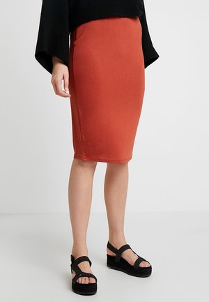 Femme portant une jupe orange ajustée au genou, un pull noir ample et des sandales à plateforme noires, debout sur un sol blanc.