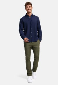 Camisa de botones azul marino, mezcla de algodón, corte relajado; combinada con pantalones verde oliva y zapatillas blancas. El modelo está de pie con las manos en los bolsillos.