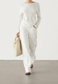 Haut blanc à manches longues, pantalon blanc texturé ample et sandales noires ; tenant un sac en toile beige tissé. Le tissu lisse contraste avec le détail côtelé.