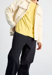 Krämfärgad fleecjacka med två bröstfickor, lagd över en ljusgul t-shirt och svarta vida byxor. Släta texturer, avslappnad passform.