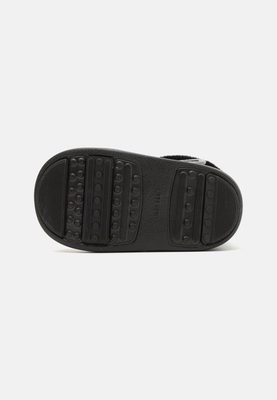 Semelle noire d'une chaussure présentant un caoutchouc texturé avec des points et des rainures surélevés pour la traction. Branding minimal visible près du talon.