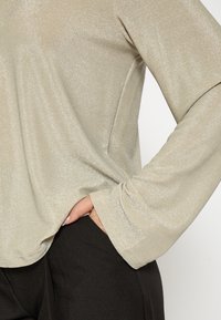 Garsa ar garām piedurknēm, izgatavota no gaiša metāliska sudraba auduma, ar brīvu piegriezumu, mīkstu tekstūru un redzamu kabatas detaļu uz melniem bikšiem.