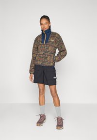 Pullover jakke med brunt mønstret stof, blå krave og orange lynlås. Kombineret med sorte shorts med to forlommer. Grå sokker og brune sko.