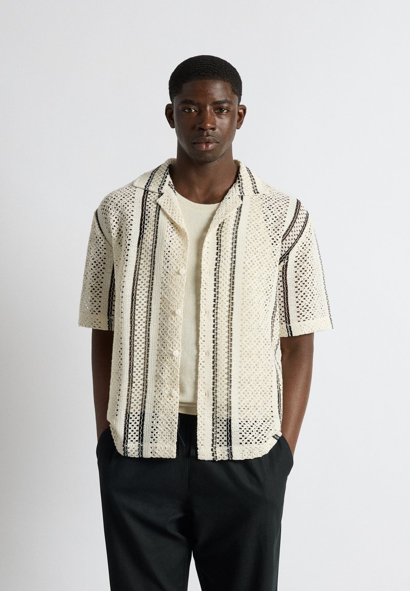 Jeune homme portant une chemise beige en crochet à manches courtes avec des rayures verticales noires sur un haut beige clair, debout les mains dans les poches d’un pantalon noir.