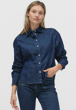 Mörkblå denimskjorta med krage, långa ärmar och silverfärgade knappar. Har en avslappnad passform och bröstficka.