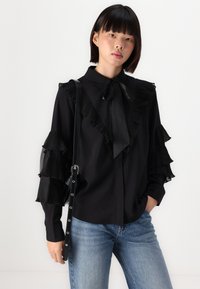 Blusa nera con dettagli plissettati a volant sulle maniche e sul colletto, con un dettaglio a cravatta e una texture liscia. Abbinata a jeans blu.
