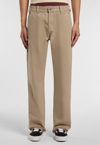 Béžové bavlněné kalhoty s rovnou nohavicí, předními kapsami a volným střihem, doplněné černými teniskami.