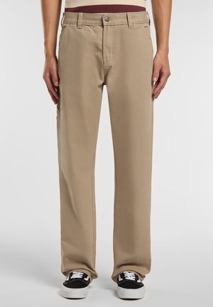 Pantaloni in cotone beige con design a gamba dritta, tasche frontali e vestibilità casual, abbinati a sneakers nere.