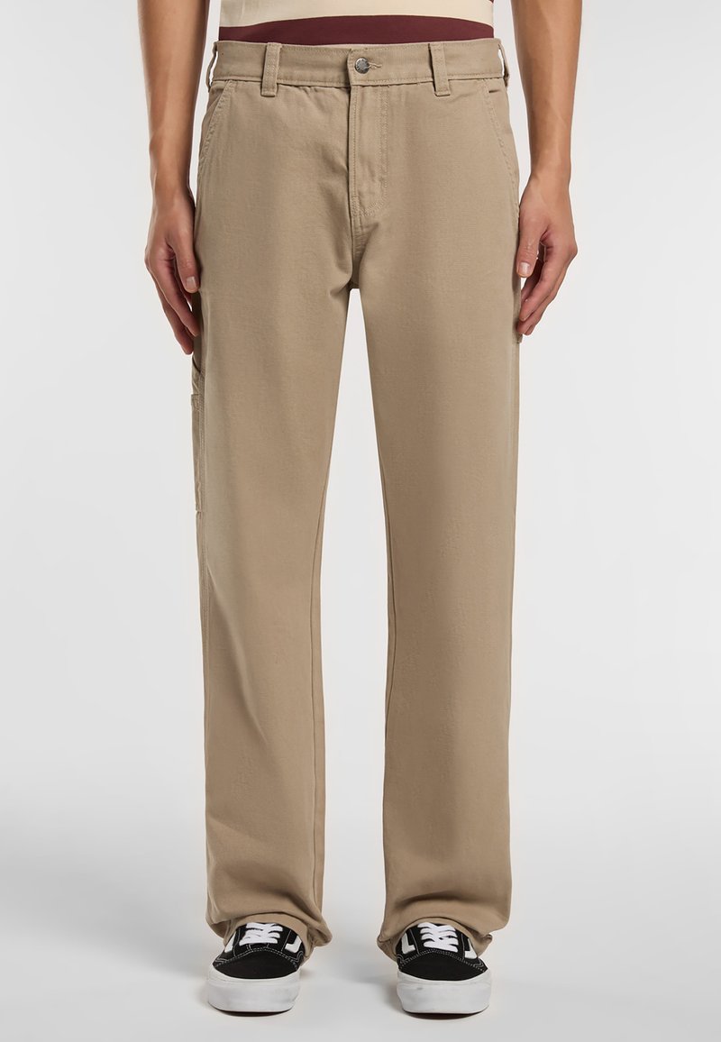 Béžové bavlněné kalhoty s rovnou nohavicí, předními kapsami a volným střihem, doplněné černými teniskami.