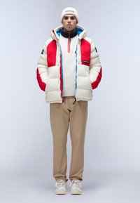 Puffjakke i creme med røde og blå detaljer, der har en lynlåsåbning foran og høj krave. Beige bukser og hvide sneakers fuldender outfittet.