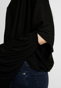 Haut noir oversize avec manches froncées, au design à enfiler et texture douce. Associé à un jean denim bleu foncé.