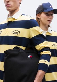 Ριγέ ράγκμπι πουκάμισα σε ναυτικό μπλε και κίτρινο με λευκό γιακά, λογότυπο στο στήθος. Μαύρη τσάντα χειρός με λογότυπο. Υφασμάτινη υφή, δομημένος σχεδιασμός.