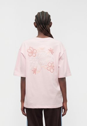 Personne avec des cheveux tressés portant un t-shirt rose clair Adidas Originals avec une broderie florale dans le dos, debout devant un fond uni.