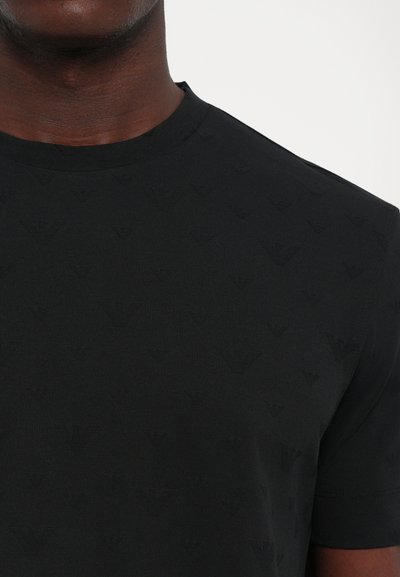 Schwarzes kurzärmliges Shirt mit dezent geprägtem, wiederholtem Logo-Muster, getragen von einer Person, die vom Hals bis zur Mitte der Brust auf einem einfarbigen Hintergrund zu sehen ist.