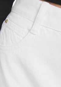 Valkoiset farkut, joissa on sileä pinta, etutasku ja ommeltu yksityiskohtia. Vyötäröltä löytyy pieni kultainen yksityiskohta.