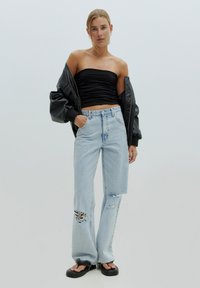 Zwarte strapless top, oversized zwarte bomberjack, lichtblauwe ripped broek met wijdere pijpen en chunky zwarte sandalen. Glad textuur en casual stijl.