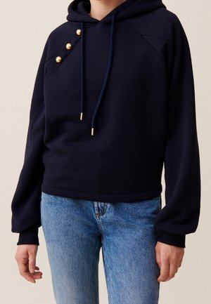 Sweat à capuche bleu marine avec des boutons dorés sur l'épaule, cordons avec embouts dorés, porté avec un jean bleu clair.