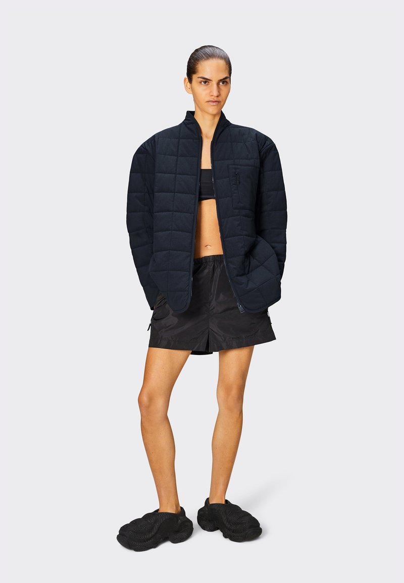 Veste noire matelassée avec col montant, fermeture éclair à l'avant et poche sur la poitrine, associée à un short noir et des chaussures texturées noires.