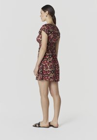 Femme debout de profil portant une robe courte à motifs rouges et noirs ainsi que des sandales noires à enfiler, sur un fond clair uni.