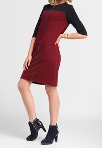 Abito bordeaux e nero con una forma aderente, maniche a tre quarti e un dettaglio con zip frontale. Abbinato a stivaletti blu navy.