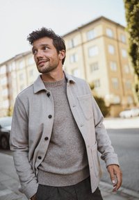 Ljusgrått ullblandat jacka med svarta knappar, en bröstficka och en stickad ljusgrå tröja under. Bakgrunden visar en urban miljö.