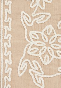 Tessuto beige con ricami floreali bianchi testurizzati; presenta design a spirale e contornati, mettendo in mostra cuciture intricate e dettagli tattili.