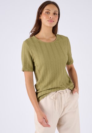 Frau trägt ein strukturiertes olivgrünes Kurzarmoberteil und hellbeige Hosen mit Kordelzug, steht mit einer Hand in der Tasche vor einem einfarbigen Hintergrund.