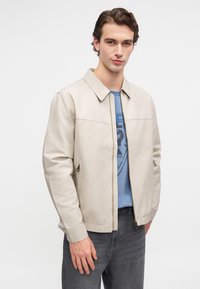 Šviesiai ruda bomberio striukė su lygiu paviršiumi, stovinčiu apykakle, priekyje su užtrauktuku ir šoniniais kišenėmis, vilkima virš mėlynos grafikės marškinėlių ir tamsių džinsų.