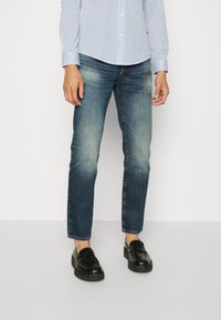 Person som bär urblekta blå jeans, svarta loafers och en vit skjorta med blå rutmönster, stående mot en enfärgad vit bakgrund.