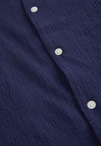 Camisa texturizada de color azul marino con tela arrugada, que cuenta con botones blancos en la parte delantera.