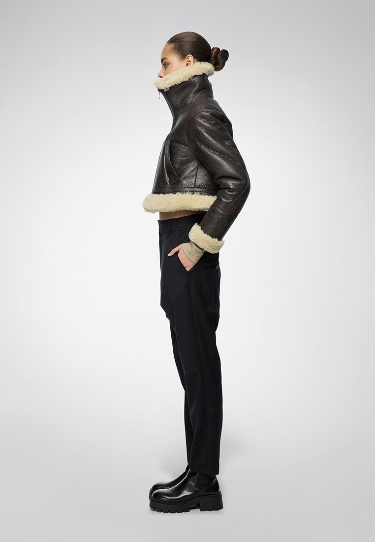 Braune Leder-Cropped-Jacke mit beigem Lammfellkragen und -manschetten, kombiniert mit schwarzen Hosen und klobigen schwarzen Stiefeln. Seitenansicht.