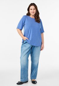Niebieska koszulka z krótkim rękawem z miękkiego materiału, połączona z jasnymi, luźnymi dżinsami. Model stoi z jedną dłonią w kieszeni.