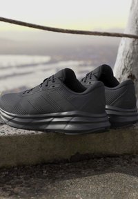 Paire de chaussures de sport noires avec tissu texturé et semelles rembourrées posées sur un rebord en pierre à l'extérieur près d'un arbre et d'une barrière en corde.