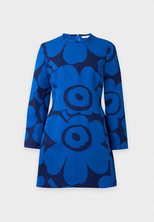 Femme portant une mini-robe bleue à manches longues avec de grands motifs floraux foncés, des chaussettes grises montant aux genoux et des ballerines bordeaux, debout avec une main dans le dos.