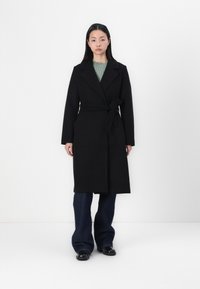 Manteau en laine noir à ceinture, avec un col châle, présentant des manches longues et un design mi-long, associé à un jean en denim foncé évasé.