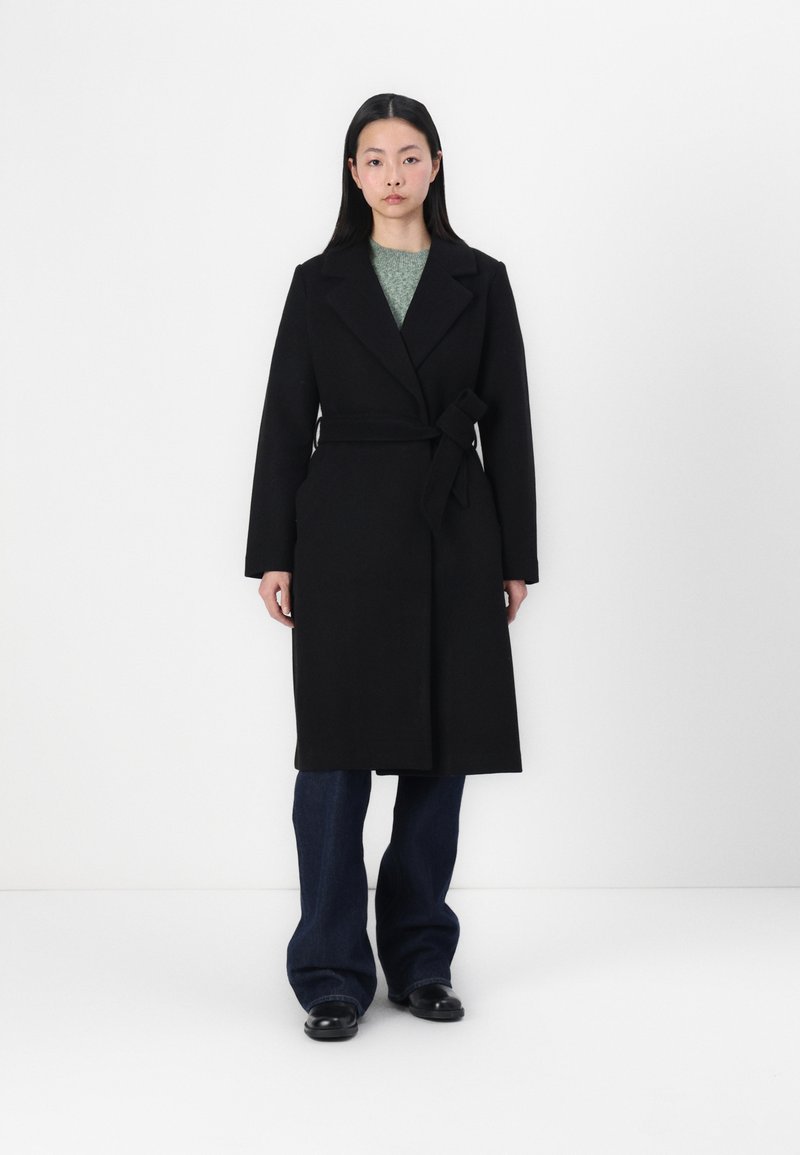 Manteau en laine noir à ceinture, avec un col châle, présentant des manches longues et un design mi-long, associé à un jean en denim foncé évasé.