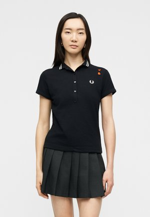 Nainen, jolla on musta lyhythihainen poolopaita, jossa on kirjailtu laakerinlehvälogo ja oranssit sydänmerkit, yhdistetty tummanharmaaseen laskoshameeseen.