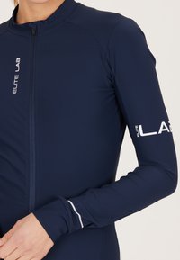 Maglia da ciclismo a maniche lunghe di colore navy con zip frontale. Le caratteristiche includono una vestibilità aderente, un tessuto strutturato e dettagli del logo stampati in bianco.