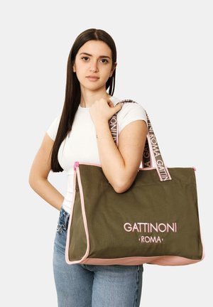 Jeune femme aux longs cheveux noirs tenant un grand sac cabas Gattinoni Roma vert olive et rose sur l'épaule, portant une chemise blanche et un jean bleu.