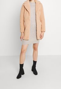 Bordeaux oversized faux shearling frakk med krave, parret med en grå strikket kjole og sorte blokhælede ankelstøvler. Glat tekstur.