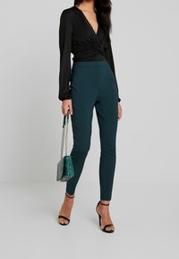 Svart långärmad topp med djup V-ringning och samlad midja, kombinerad med turkosa figurnära byxor. Accessoarer i form av en turkos handväska och svarta klackar.