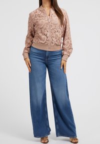 Veste bomber rose avec une texture en dentelle florale complexe, poignets et ourlet côtelés. Associée à un jean bleu à jambes larges et des sandales marron à brides.