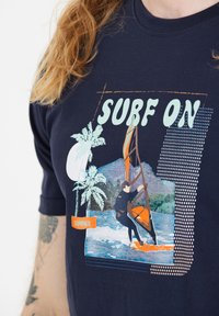 Laivansininen t-paita, jossa on graafinen printti purjelautailijasta, palmupuista ja täysikuusta, sekä tekstikorostukset "SURF ON" ja "SUMMER".