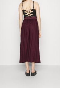 Femme portant un débardeur noir à lacets dans le dos, une jupe midi violette foncée taille haute et des chaussures à plateforme noires à enfiler, se tenant dos au spectateur.