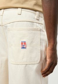 Los pantalones de mezclilla beige Wrangler cuentan con un bolsillo trasero con un parche de logo en rojo y azul, costuras en contraste y una textura suave.
