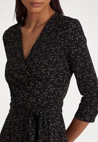 Svart omslagsklänning med små gyllene blommönster, V-ringning, tre fjärdedelsärmar och knytning i midjan. Slät tygtextur.