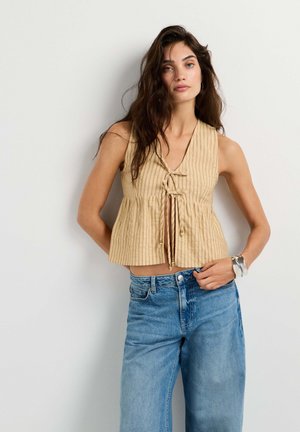 Vrouw met lang donker haar draagt een beige mouwloos gestreept topje met voorstrikjes en losse blauwe jeans, staand tegen een effen lichte achtergrond.